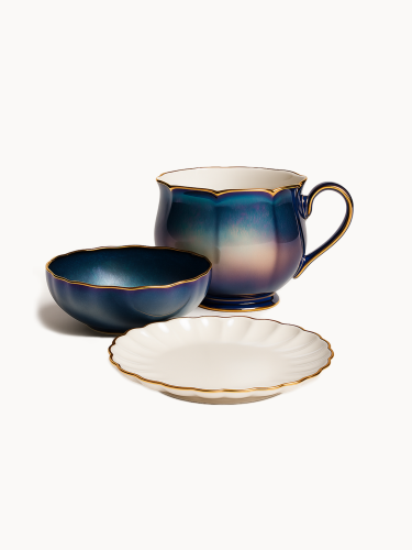 Elegancki ceramiczny zestaw śniadaniowy zawiera duży kubek oraz pasującą miseczkę z iryzującym, niebiesko-fioletowym szkliwem i falowanymi, złotymi rantami. Na pierwszym planie znajduje się biały, karbowany spodek ze złotym brzegiem.