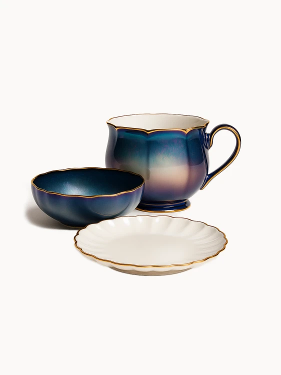 Elegancki ceramiczny zestaw śniadaniowy zawiera duży kubek oraz pasującą miseczkę z iryzującym, niebiesko-fioletowym szkliwem i falowanymi, złotymi rantami. Na pierwszym planie znajduje się biały, karbowany spodek ze złotym brzegiem.