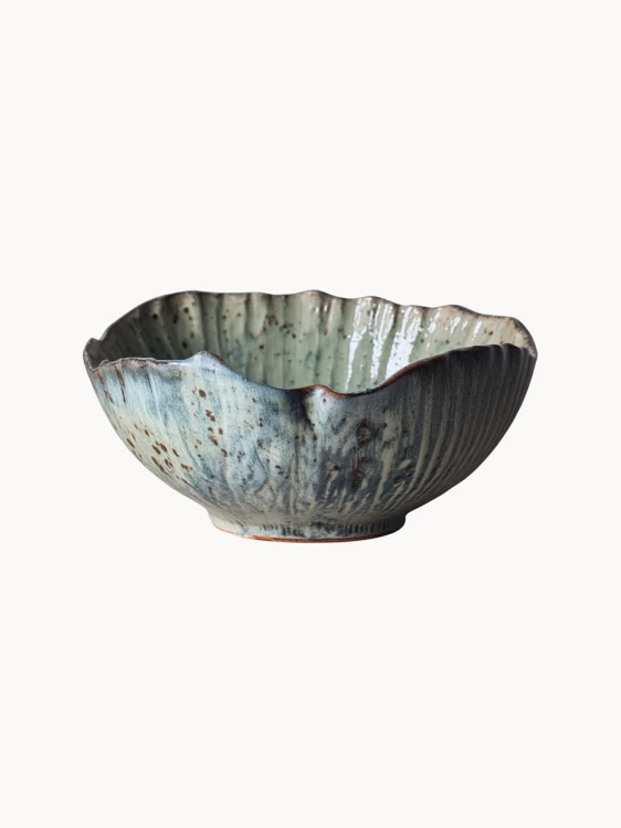 Rustykalna ceramiczna miska pokryta melanżowym jasnoniebieskim i beżowym szkliwem z brązowymi, nakrapianymi detalami na jednolitym białym tle.