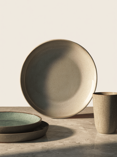 Ceramiczny talerz o nakrapianej, jasnoszarej i beżowej strukturze z lekko błyszczącym wykończeniem w otoczeniu pasujących talerzy i kubków.