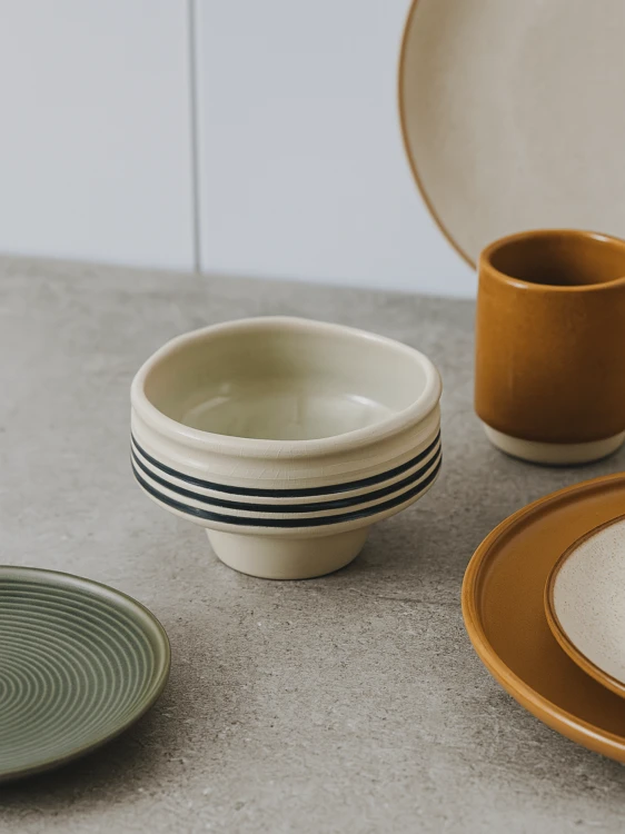 Kremowa ceramiczna miska na podwyższeniu z czarnymi, poziomymi paskami stoi na szarym blacie. Minimalistyczna aranżacja naczyń obejmuje talerze w kolorach ziemi oraz ochrowy kubek w tle.