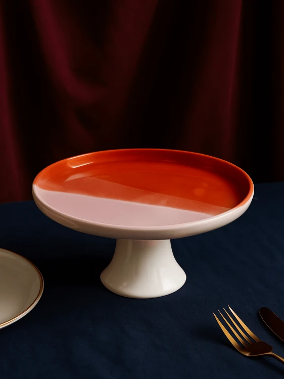 Ceramiczna patera z dwukolorowym blatem na ciemnoniebieskim obrusie. Gładka powierzchnia dzieli się na błyszczący pomarańcz i matowy jasny róż, zaprezentowane obok złotego widelca i fragmentu talerza.